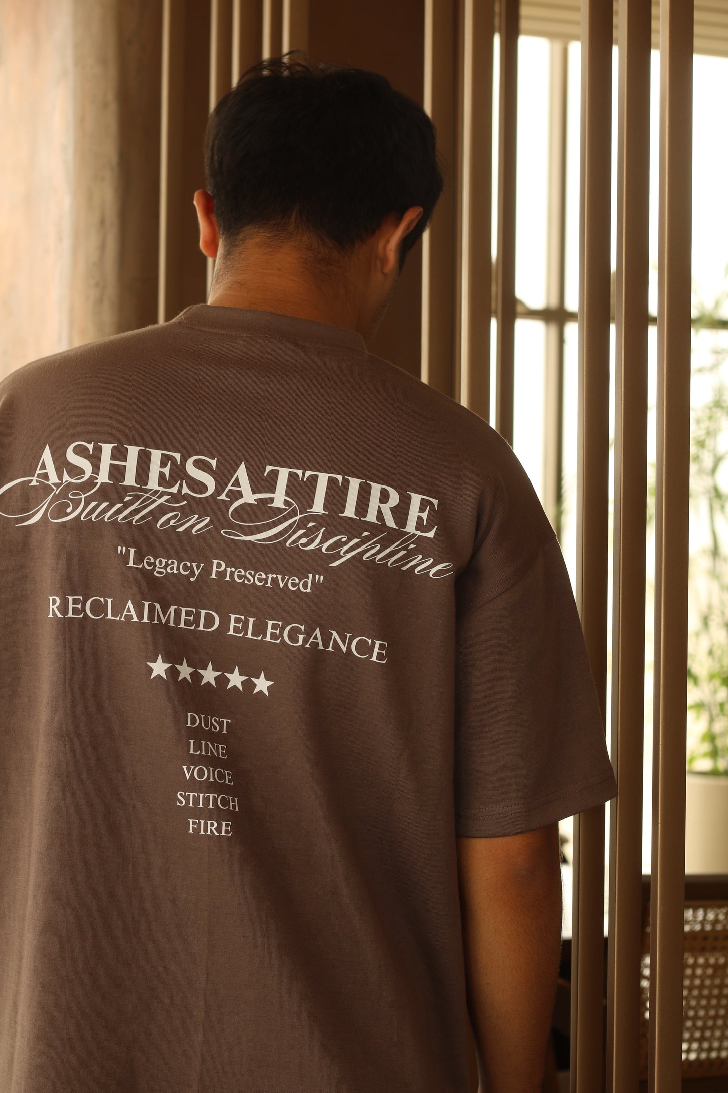 Person wearing a brown shirt with text, standing in front of horizontal blinds.